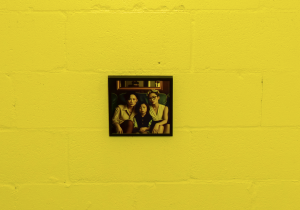 Image: Lovers’ Portraits, a small square image by Linye Jiang showing a queer Asian family, hangs on a bright yellow brick wall. Photo by Ji Yang.